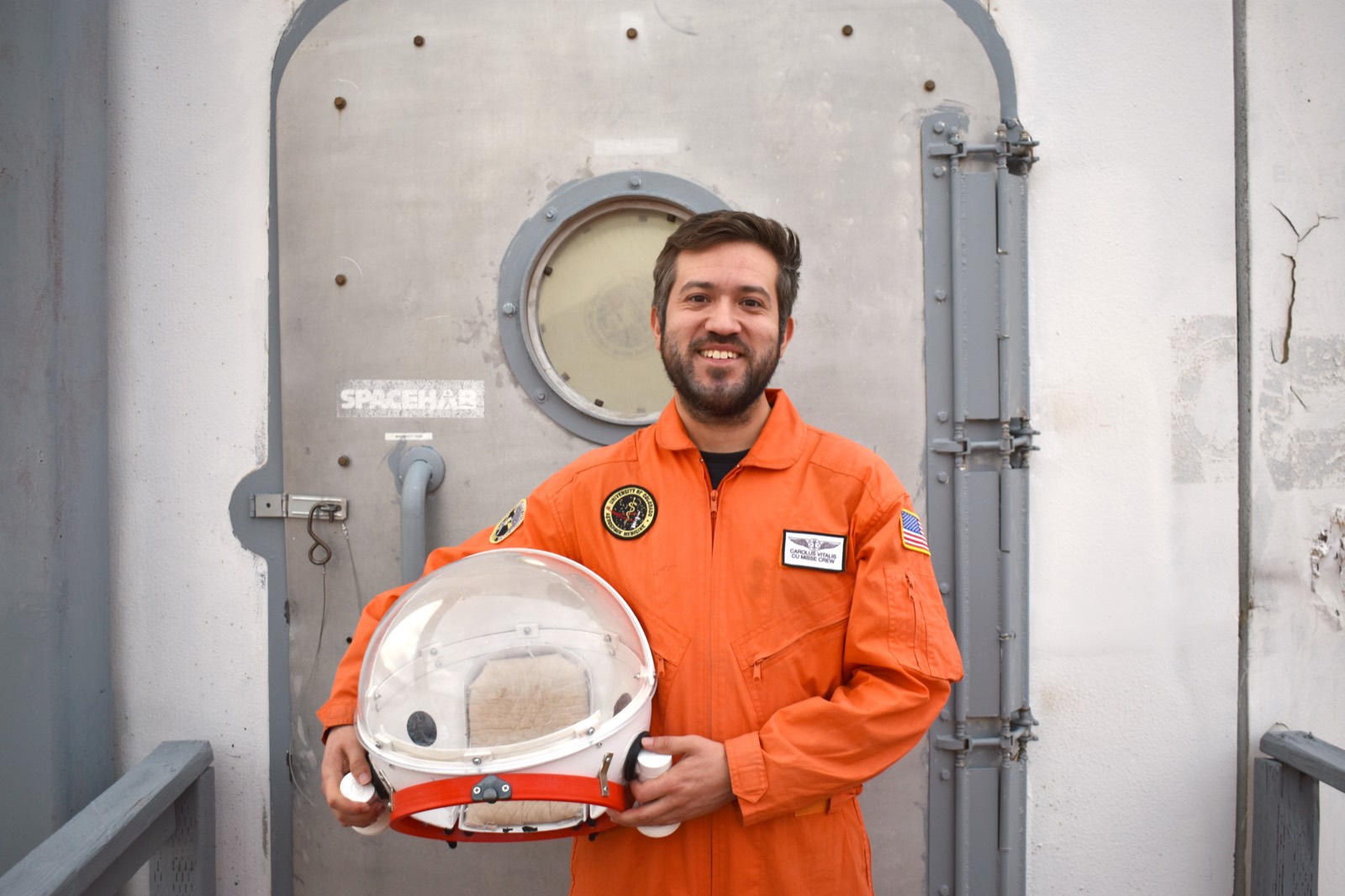 Carolus Vitalis in an orange flight suit, holding an analog space helmet at the airlock of the Mars Desert Research Station habitat.